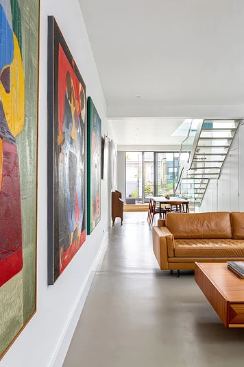 Bright hallway with colorful framed artwork leading to a living room with a tan leather sofa and a staircase